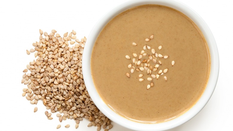 A white bowl of homemade tahini beside a pile of sesame seeds 