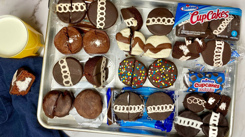 A tray with 11 different types of creme filled chocolate cupcakes on it and a glass of milk for taste testing