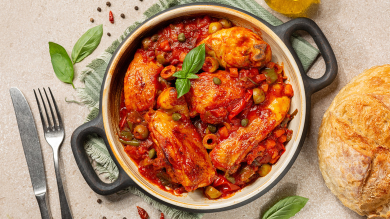 Black pot with chicken cacciatore on a countertop with break and silverware next to it