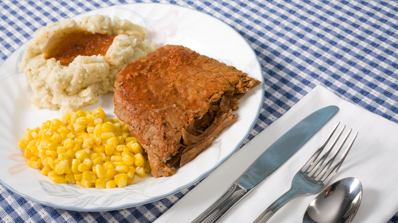 Plate of swiss steak, mashed potatoes with gravy, and corn sitting on a blue checkered table