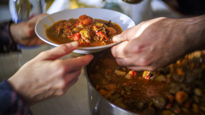 A pair of hands giving a white bowl of venison stew to another person