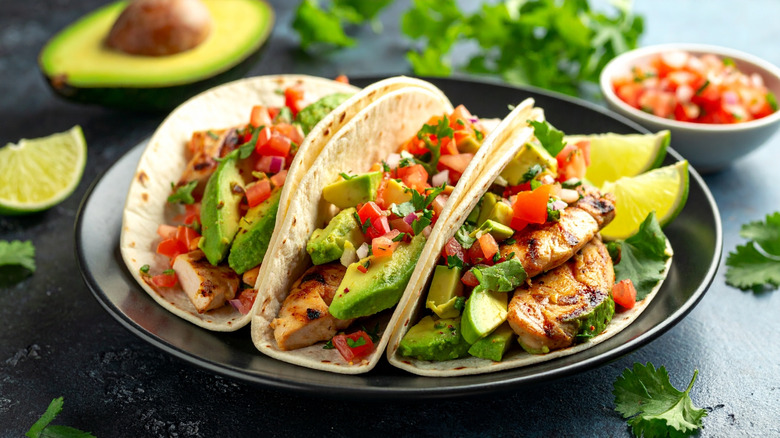 A plate of tacos with chicken, avocados, tomatoes, basil, pico de gallo, and slices of lime