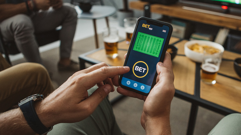 A man's hands holding a mobile phone and betting while drinking with friends.