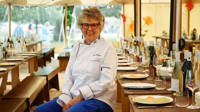 Prue Leith in her chef's outfit, sitting at a long table set inside a tent