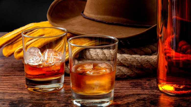 Two glasses of bourbon withh ice next to bottle, cowboy hat, and rope