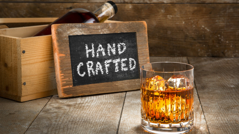 Glass of bourbon with ice next to bottle in crate with sign reading "hand crafted"