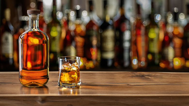 Simple bottle of bourbon next to glass of bourbon with ice on bar counter with blurred bottles in background
