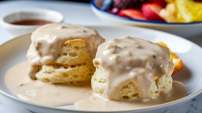 A close-up of biscuits and gravy on a plate with a bowl of fruit visible in the background