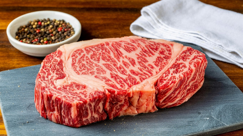 Large piece of wagyu beef on gray slate with peppercorns and towel in background