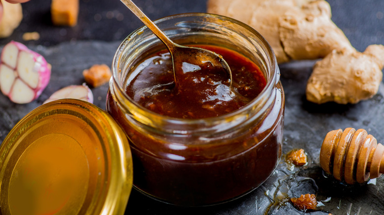 A jar of marinade with a spoon and fresh ginger in background