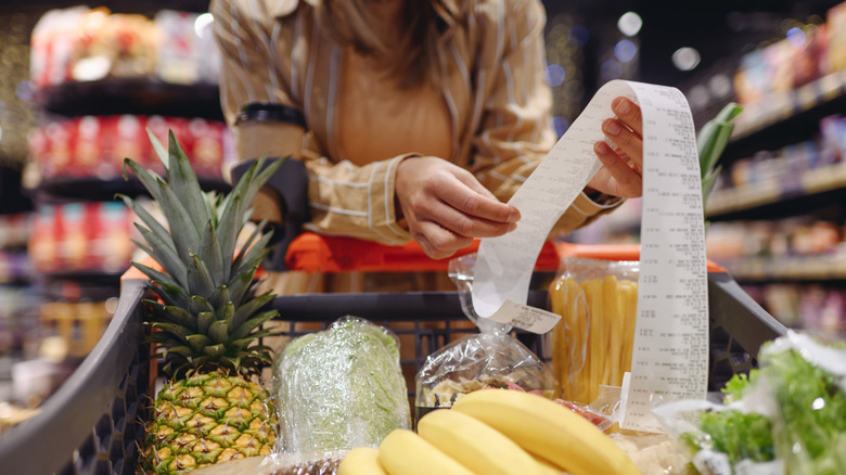 A woman leaning into her grocery cart looking at a long receipt.