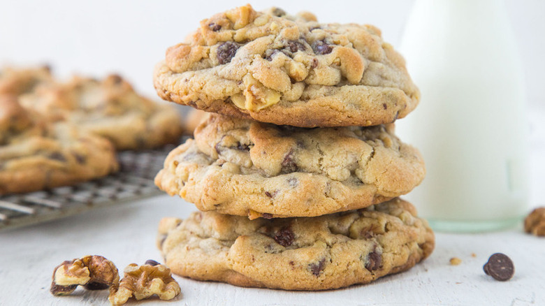 Three large chocolate chip cookies in pile next to cooling rack of more cookies and pitcher of milk