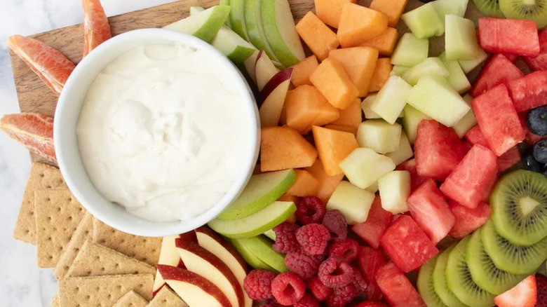 Marshallow fruit dip in ramekin on board filled with cut fruits, melons, berries, and graham crackers