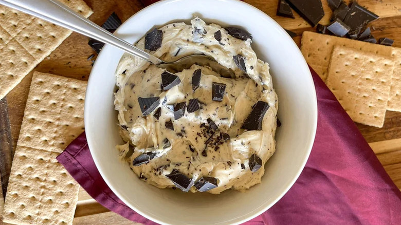 bowl of cookie dough dip with spoon topped with chocolate chunks surrounded by graham crackers and chocolate on table