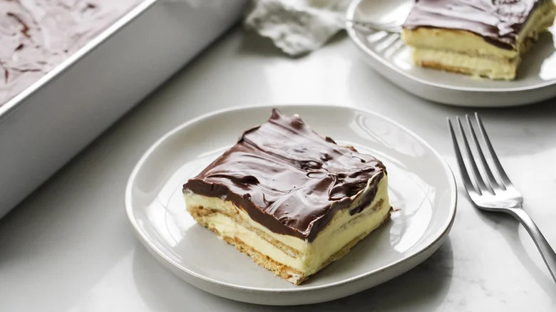 Slice of no-bake chocolate eclair cake on plate next to fork, another plate with a slice, and ceramic bakeware with the dessert