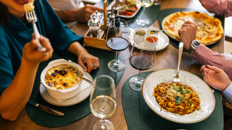Four people eating various dishes at Italian restaurant, like pasta and pizza, with glasses of red and white wine