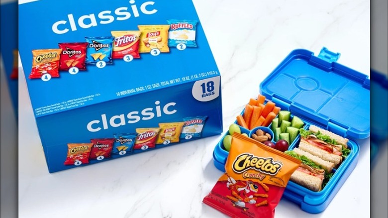 Package of Frito Lay chip bags containing Cheetos, Doritos, Fritos, Lay's Classic, and Ruffles with lunch box of veggies and sandwich halves.