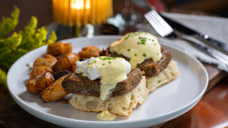 eggs benedicts with scrapple on a white plate, a candle glowing in the background