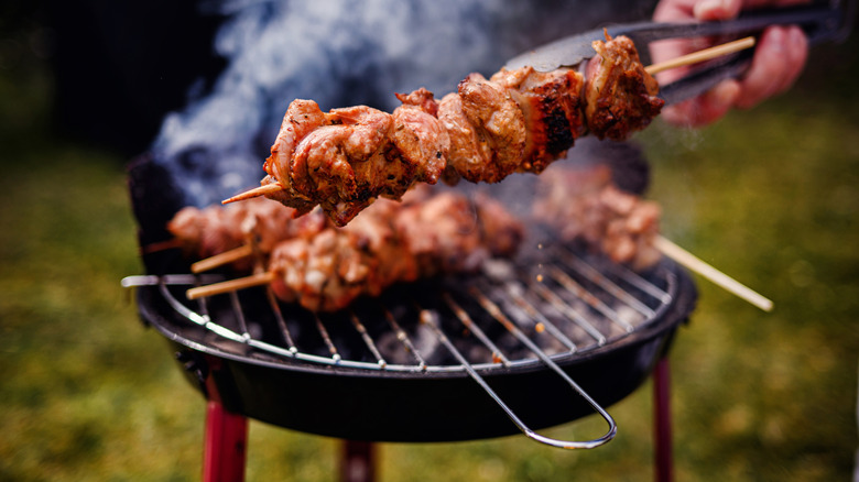 Hand holding a skewer of meat with charcoal grill in background
