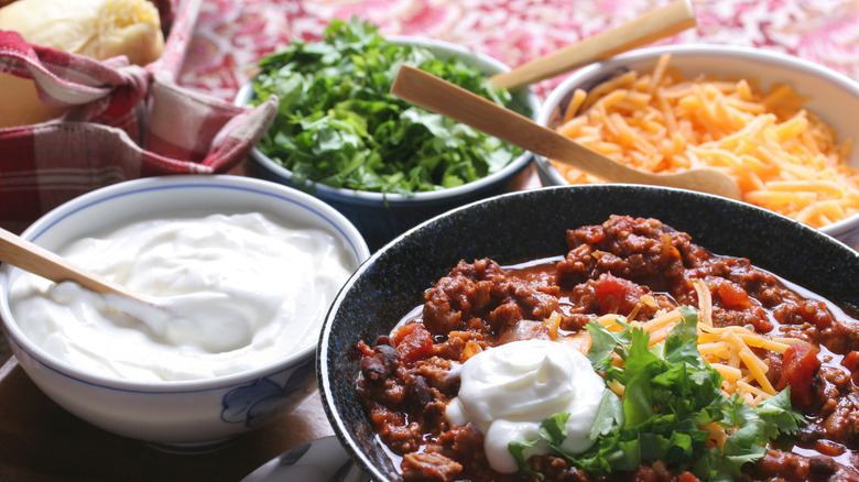 A bowl of chili loaded with sour cream, shredded cheese, and fresh herbs next to bowls of sour cream, shredded cheese, herbs, and bread rolls