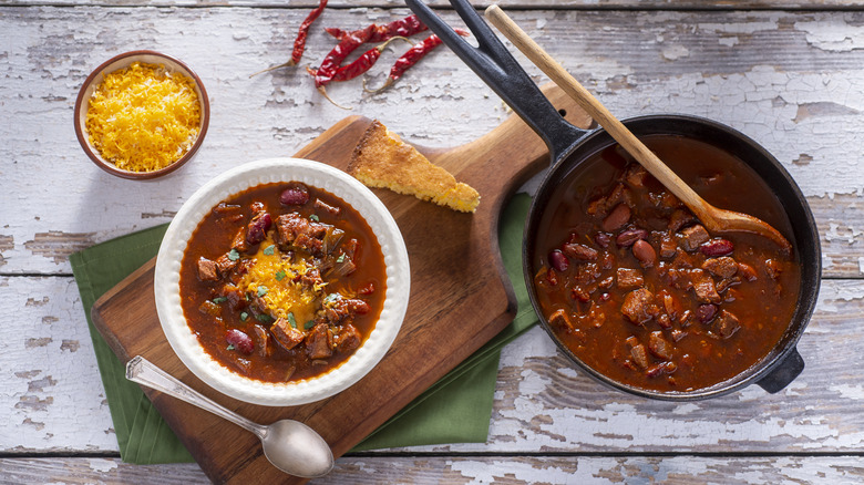 A pot and bowl of chili topped with shredded cheese next to a bowl of shredded cheese and peppers
