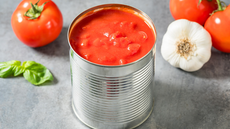 An open can of diced tomato next to fresh tomato, garlic, and basil