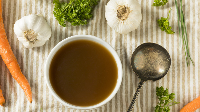 A bowl of beef broth next to fresh carrots, garlic, and herbs