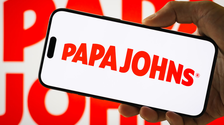 A person holding a smartphone with the Papa Johns logo on it in front of a Papa Johns logo