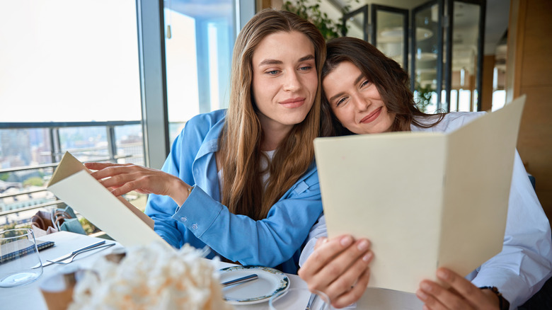 Two women in a restaurant reading a menu, one resting her head on the other's shoulder