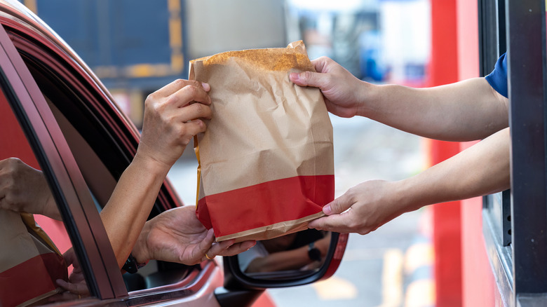 A person grabbing a bag of food out of a drive thru window from their red car