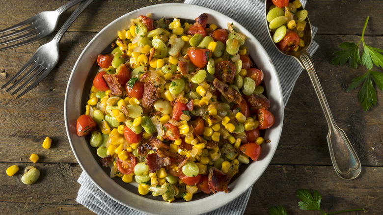 Succotash with bacon and tomatoes in a bowl, placed on top of a table napkin along with a spoon and two forks
