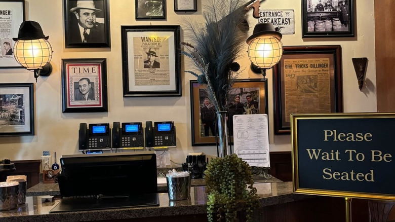 The front desk at Exchequer Restaurant showing a sign to wait to be seated, picture of Al Capone, and a speakeasy sign