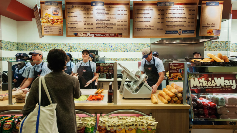 A customer ordering at Jersey Mike's while employees make sandwiches.