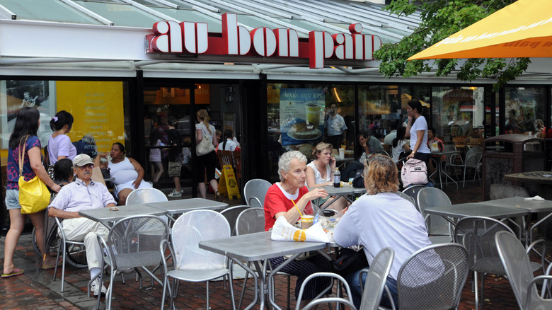 Diners eat on patio tables in front of an entrance to Au Bon Pain