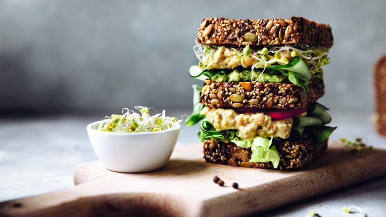 Sandwich stacked on wooden board with bowl of sprouts