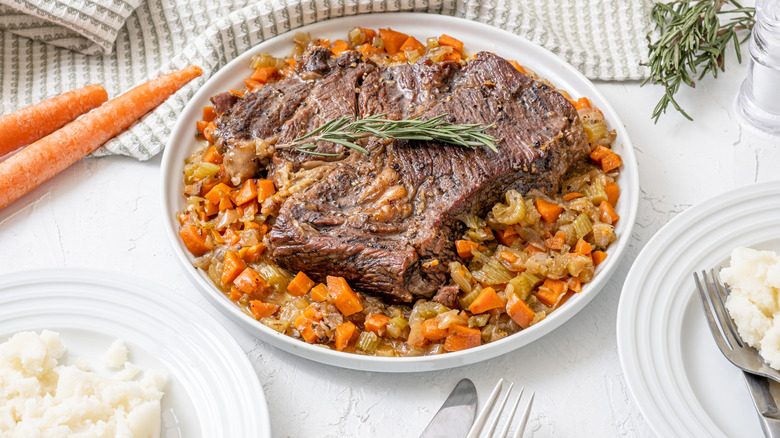 A plate of pot roast next to some raw carrots and plates with mashed potatoes