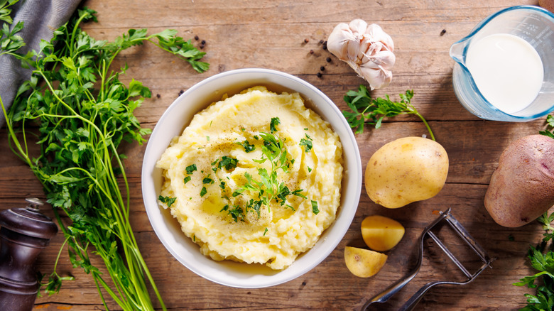 A bowl of mashed potatoes next to raw potato and garlic, milk, a peeler, and fresh herbs