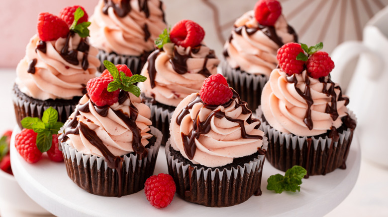 Plate of chocolate cupcakes with pink frosting and a a ganache drizzle, fresh raspberries, and mint sprigs