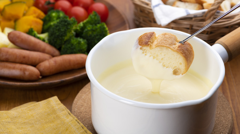Bread being dipped into a pot of cheese fondue, veggies on plate and bread basket in background.