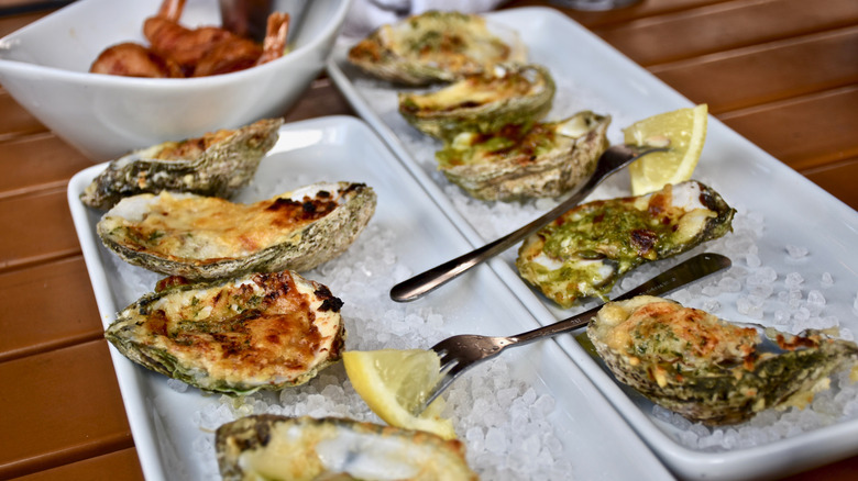 Two white rectangular serving trays topped with oysters Rockefeller on rock salt, with lemon wedges.