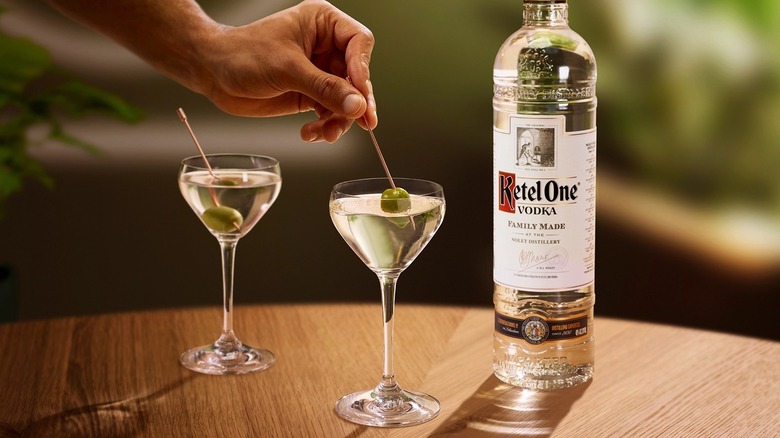 A bottle of Ketel One vodka next to two dirty olive martinis