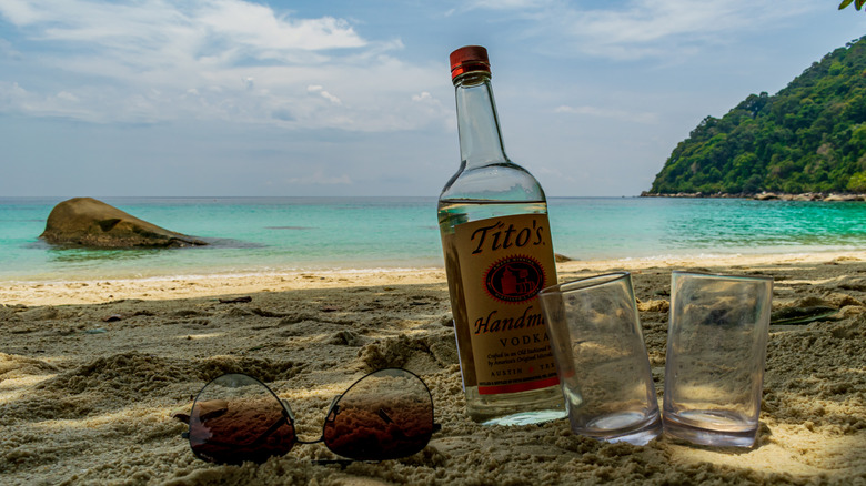 A bottle of Tito's vodka with two empty glasses and sunglasses in the sand on a beach