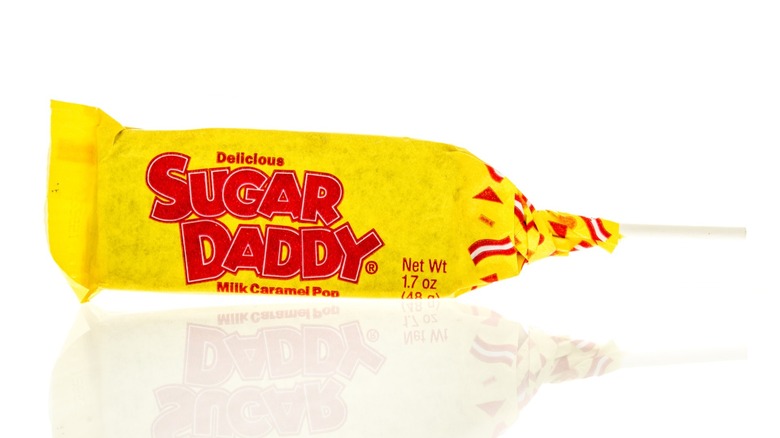 A Sugar Daddy candy in wrapper with stick on white background with reflection.