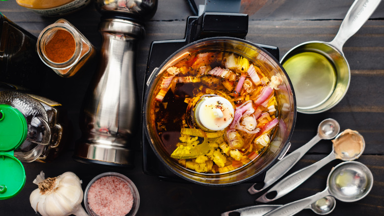 Italian ingredients in a food processor, including onions, artichokes, and peppers, surrounded by measuring cups and spoons, salt, pepper, garlic, and spices