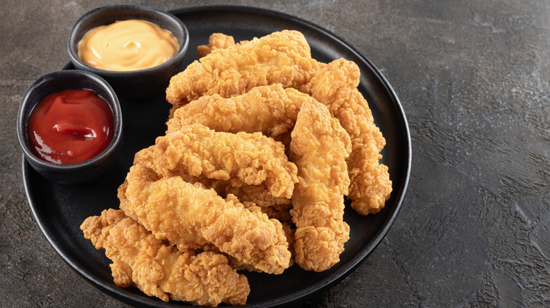 A black plate of crispy chicken strips with ketchup and another dipping sauce in black ramekins
