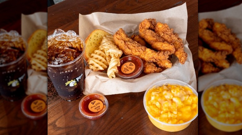 A basket of PDQ (People Dedicated to Quality) chicken tenders with crisscut fries and a piece of bread, a side of mac and cheese, a soda, and various dipping sauces