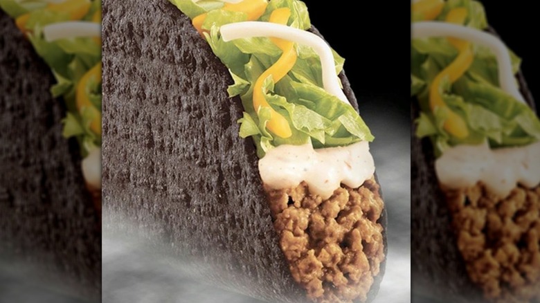Taco Bell taco with a hard black shell, topped with ground beef, jack cheese, lettuce, and Baja sauce.