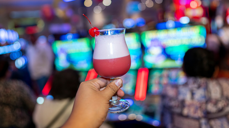 Hand holding a pink and white cocktail in front of slot machines