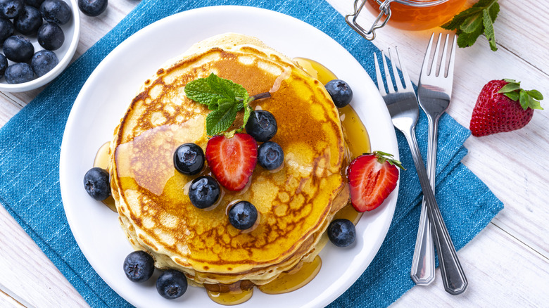 A stack of pancakes with fresh fruit and syrup on a white plate with two forks alongside it