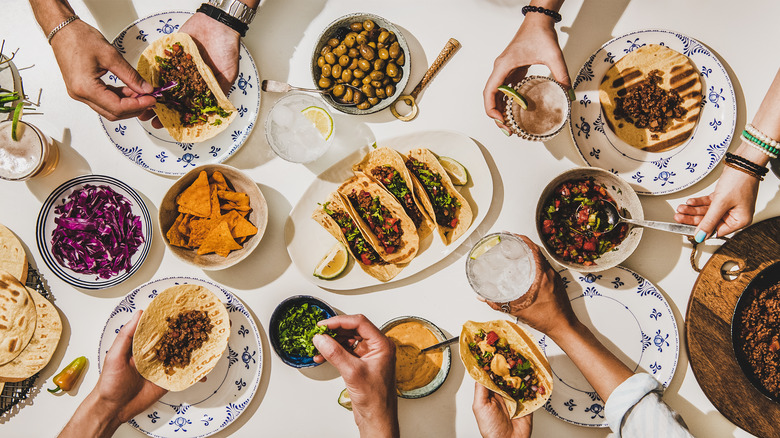 a festive dinner scene with plates of tacos and toppings and hands reaching in to make combinations